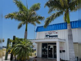 A view of a fishing and diving center surrounded by palm trees, showcasing a sunny day atmosphere. The building features a sign indicating its services.
