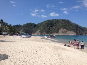 A vibrant beach scene with groups of people enjoying the sun and water, surrounded by lush hills and boats in the distance — a perfect destination for relaxation and fun.