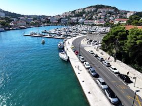 Aerial view of a marina with various boats docked and a road lined with parked cars, showcasing a vibrant coastal scene.