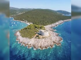 A stunning aerial view of a rocky coastline with a historic structure on the shore, surrounded by crystal-clear waters and lush greenery — a picturesque destination for boaters and nature lovers.