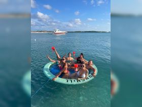 A lively group enjoys cocktails while relaxing on a floating mat in clear waters, perfect for a fun day out on the water.
