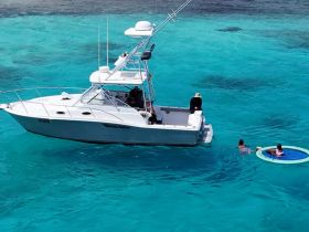 A group enjoys a fun day on the water, with a boat anchored nearby and swimmers relaxing on a floating mat in the vibrant turquoise sea.
