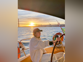 A woman steers a sailboat during a stunning sunset, with a picturesque bridge in the background — an ideal moment for sailing enthusiasts and sunset lovers.