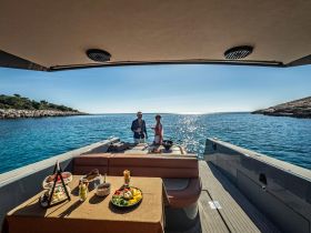 A couple enjoys a delightful spread of food and drinks on the deck of a boat, surrounded by stunning coastal scenery — perfect for a relaxing day on the water.