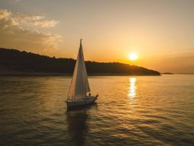 A stunning aerial view of a sailboat gracefully sailing at sunset, reflecting golden hues on the calm waters — an ideal scene for sailing enthusiasts and sunset lovers.