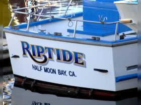 A close-up view of the 'Riptide' boat docked in Half Moon Bay, CA, showcasing its vibrant blue and white design.