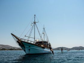 A lively scene featuring guests enjoying water activities around a beautiful boat, with one person diving off the side and others paddleboarding — perfect for summer adventures on the water.