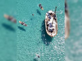 A vibrant scene of guests enjoying a day on the water, featuring paddleboarding and swimming in crystal-clear turquoise waters, with a charming boat in the backdrop.