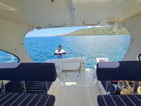Inside view of a boat's cabin featuring seating and a helm, with a crew member preparing for the journey on calm blue waters.