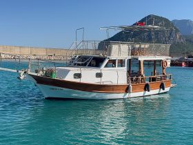 A beautifully crafted boat named 'Deniz Can' docked in clear turquoise waters, showcasing its elegant design against a stunning mountainous backdrop.