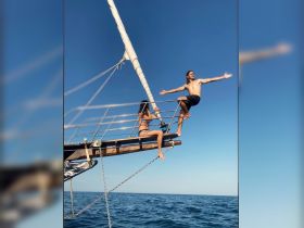 A couple enjoys an adventurous moment on the bow of a sailboat, embracing the ocean breeze and sunshine — perfect for memorable sailing experiences.