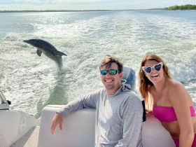 A playful dolphin leaps out of the water alongside a couple enjoying their boat ride, capturing a joyful wildlife moment in the open sea.