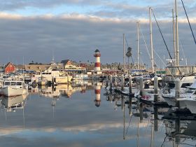 A serene view of a marina filled with various boats, reflecting beautifully in the calm waters, with a charming lighthouse in the background.