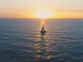 A stunning aerial view of a sailboat gliding across the ocean at sunset, creating a picturesque scene perfect for sailing enthusiasts and sunset lovers.