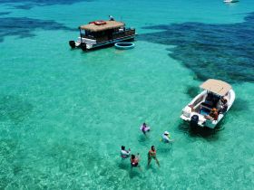 A lively group enjoys a fun day in the clear turquoise waters, swimming and interacting near their boats — a perfect sandbar stop for relaxation and enjoyment.
