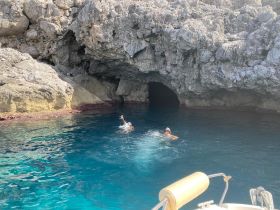 Two swimmers enjoy the crystal-clear waters near a rocky cave, showcasing a stunning natural landscape perfect for adventure seekers.