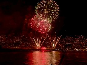 A stunning display of fireworks lights up the night sky over a coastal city, creating a vibrant backdrop for celebrations on the water.