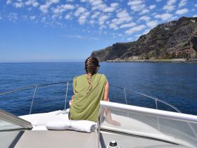 A woman enjoys the serene ocean view from the bow of a boat, with stunning coastal cliffs in the background — a perfect moment for relaxation and sightseeing.