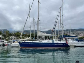 A beautiful Oceanis 411 sailboat docked in a marina, showcasing its elegant design against a backdrop of lush hills and cloudy skies.