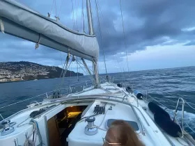 A scenic view of a sailboat navigating through the ocean, showcasing the vessel's structure against a backdrop of coastal hills and a dramatic sky.