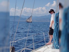 A person enjoys a scenic sailing experience on a clear day, with a beautiful backdrop of mountains and another sailboat in the distance.
