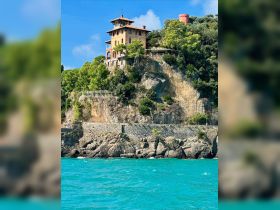 A stunning view of a coastal villa perched on a cliff, surrounded by lush greenery and turquoise waters — a picturesque landmark perfect for scenic boat tours.