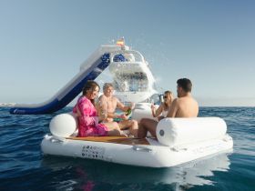 A lively group enjoys drinks and splashes on a floating mat near a yacht with a water slide, capturing the essence of summer fun on the water.