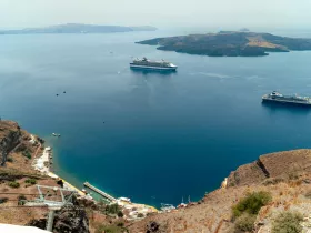 A stunning aerial view of a serene harbor in Santorini, featuring multiple cruise ships anchored in the calm blue waters, surrounded by picturesque islands and rugged coastline.