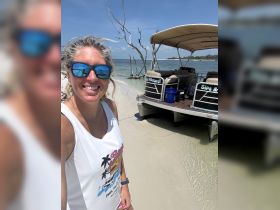 A joyful selfie by the beach with a pontoon boat in the background, showcasing a perfect day for fun and relaxation on the water.