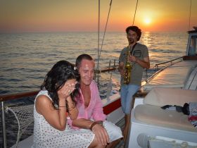 A couple enjoys a romantic moment aboard a boat at sunset, accompanied by a saxophonist, creating a perfect atmosphere for celebration.