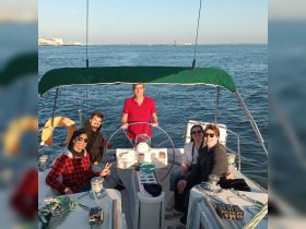 A cheerful group enjoys a sailing experience on a boat, with one person at the helm and others relaxing and celebrating — perfect for memorable outings on the water.