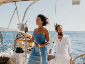 A couple enjoys a serene sailing experience on the open ocean, with one at the helm and the other admiring the view — perfect for romantic getaways or sailing adventures.