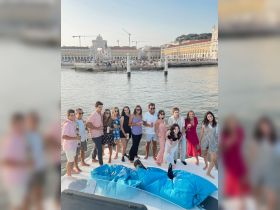 A lively group of friends enjoys a festive moment aboard a boat, celebrating with drinks and laughter against the backdrop of a beautiful waterfront in Lisbon.