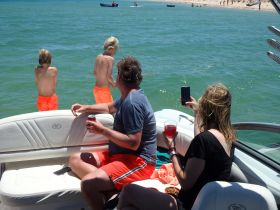 A family enjoys a sunny day on the water, with kids preparing to jump in while adults relax on the boat with drinks — a perfect summer outing.