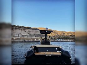 A striking view of a sleek boat from the back, anchored near a scenic shoreline, showcasing its elegant design against a backdrop of hills and clear skies.