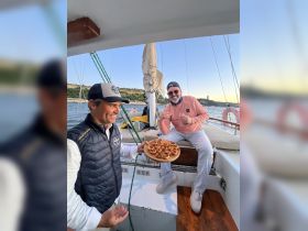 Two friends enjoy a delightful culinary experience aboard a sailboat, sharing delicious appetizers and drinks while sailing — perfect for memorable outings on the water.