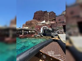 A stunning view from a boat showcasing a vibrant restaurant along the coastline, surrounded by clear turquoise waters and rocky cliffs — perfect for a scenic dining experience.