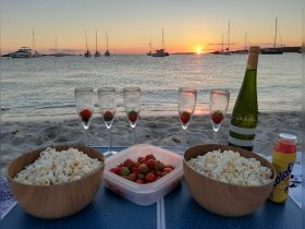 A picturesque beach setup featuring bowls of popcorn and strawberries, champagne glasses, and a bottle of wine, all set against a stunning sunset backdrop — perfect for a romantic evening or a special celebration.