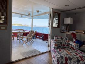 A cozy interior view of a boat's cabin, showcasing a dining area with a table and chairs, leading out to a scenic deck with stunning water views.