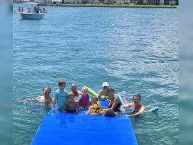 A joyful group of adults and children enjoy a fun day in the water, playing on a floating mat and swimming together — perfect for family outings and summer adventures.