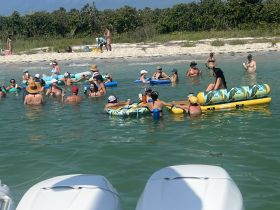 A lively group enjoys a sandbar stop, relaxing and socializing in the water with drinks and inflatable floats — the perfect setting for a fun day on the water.