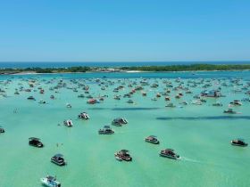A vibrant scene of numerous boats gathered at a sandbar, showcasing a lively day on the water with people enjoying the sun and swimming in the turquoise shallows.