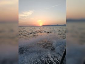 A serene sunset view over the water, captured from a moving boat, showcasing the tranquil beauty of the sea and distant mountains.