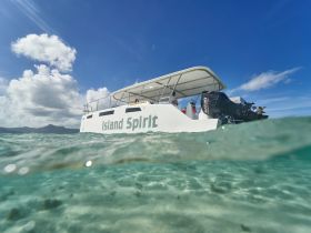 A stunning view of the 'Island Spirit' catamaran cruising through crystal-clear waters, showcasing its sleek design against a backdrop of blue skies and distant mountains.