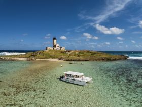 A picturesque view of a catamaran anchored near a scenic island with a historic lighthouse, showcasing a perfect day for swimming and exploration in crystal-clear waters.