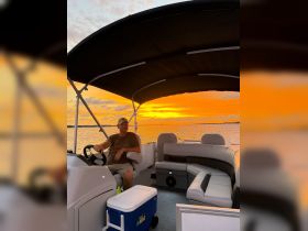 A serene sunset view from inside a pontoon boat, featuring a man at the helm, showcasing comfortable seating and a cooler — perfect for relaxing on the water.