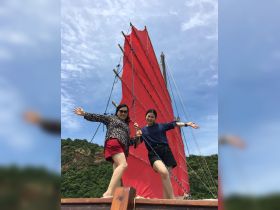 Two friends joyfully pose on the deck of a boat with vibrant red sails, capturing a moment of fun and adventure on the water.