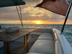 A serene sunset view from the deck of a boat, showcasing a stylish seating area and a beautiful horizon over the water.