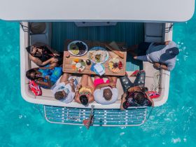 A lively group enjoys a delicious spread of food and drinks aboard a pontoon, surrounded by stunning turquoise waters — perfect for a fun day on the water.
