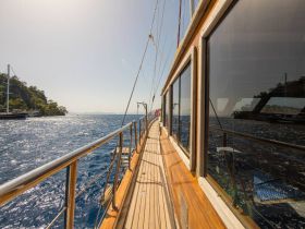 A stunning view along the side of a yacht cruising through sparkling blue waters, showcasing the vessel's elegant design and the beautiful coastal scenery.